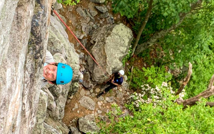 a student in the foreground smiles up at the camera while a person belaying looks on from below